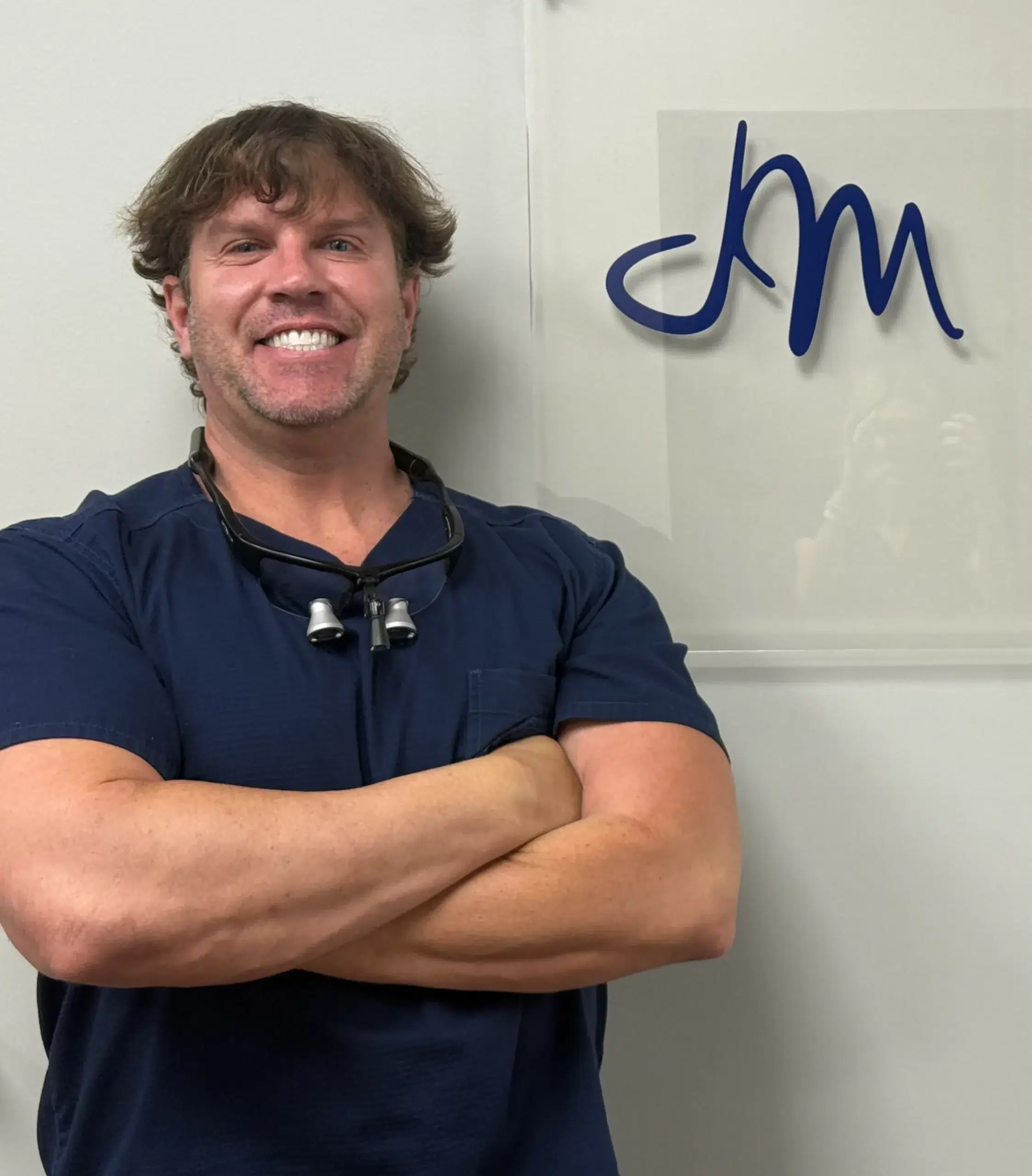 Dr. Jay Measells in navy scrubs and dental loupes, smiling with arms crossed beside the practice logo